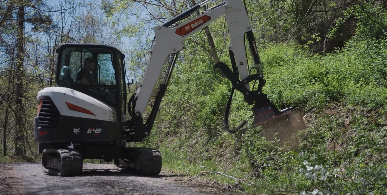 Compact excavator with brush hog attachment clearing dense vegetation and saplings from wooded terrain