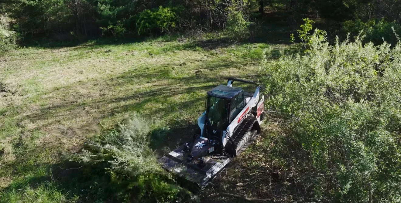 Skid steer loader with brush hog attachment clearing overgrown vegetation from wooded lot
