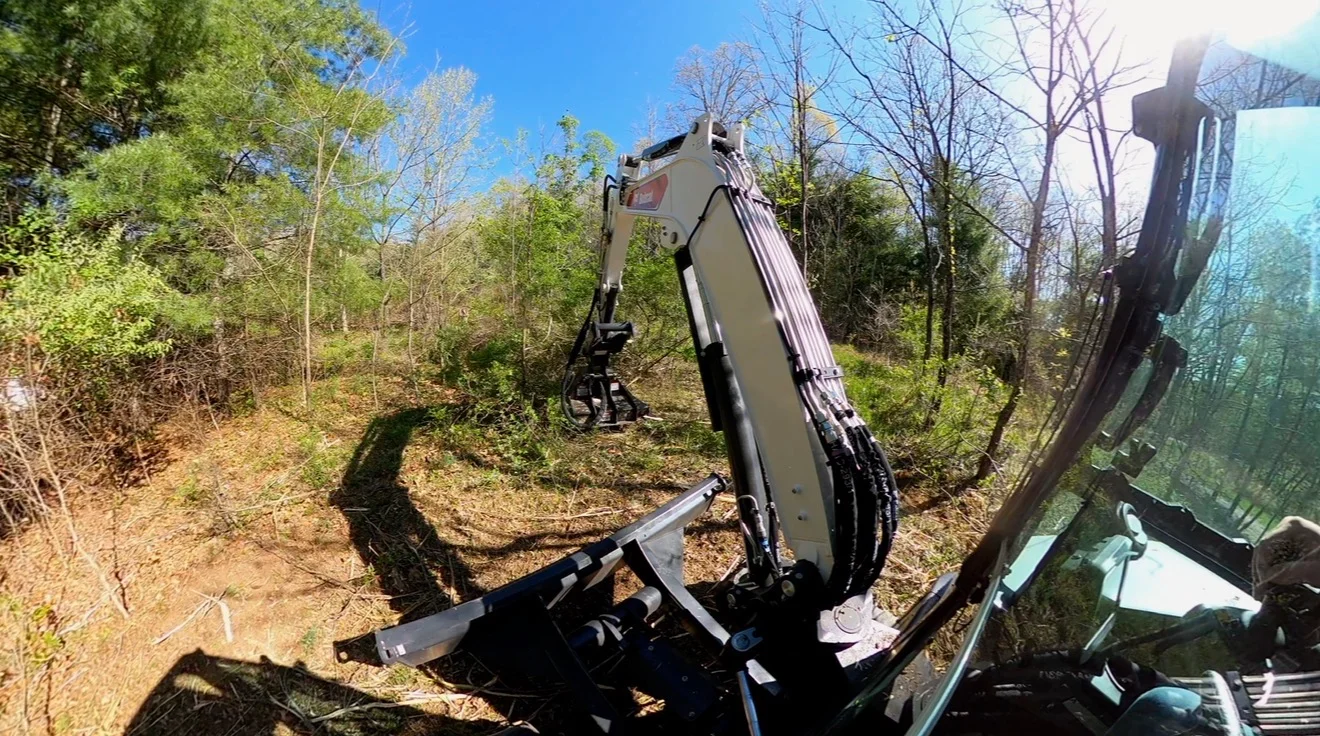 Excavator with brush hog attachment clearing overgrown vegetation and small trees from wooded slope