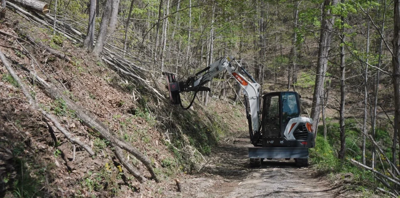 Compact excavator with brush hog attachment clearing dense vegetation on wooded hillside