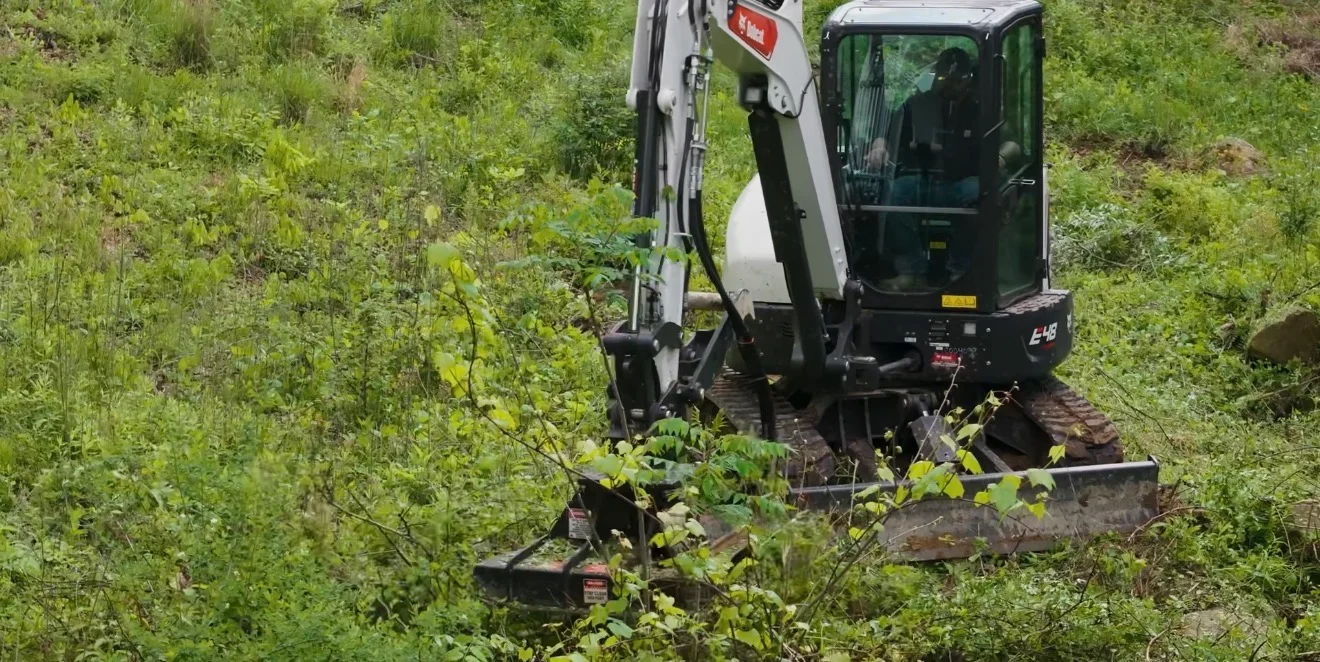 Compact excavator with rotary cutter attachment clearing dense brush and vegetation from sloped terrain